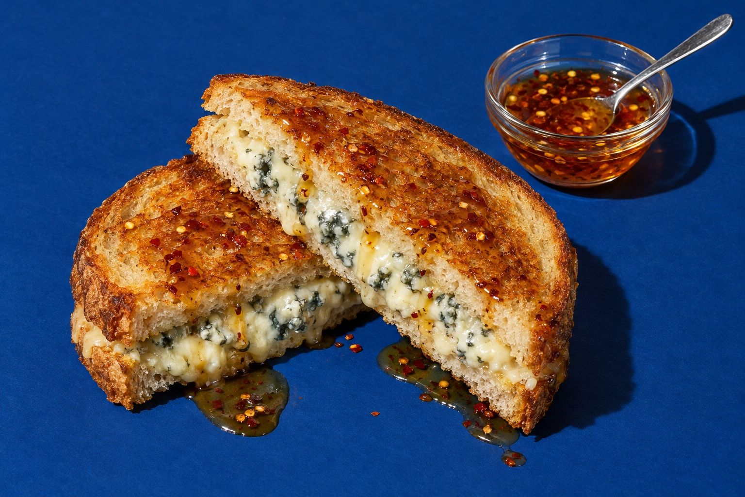 En delad toast med smält blåmögelost i snittytan och chilihonung ringlad över den gyllene ytan, bredvid en liten glasskål med chilihonung.