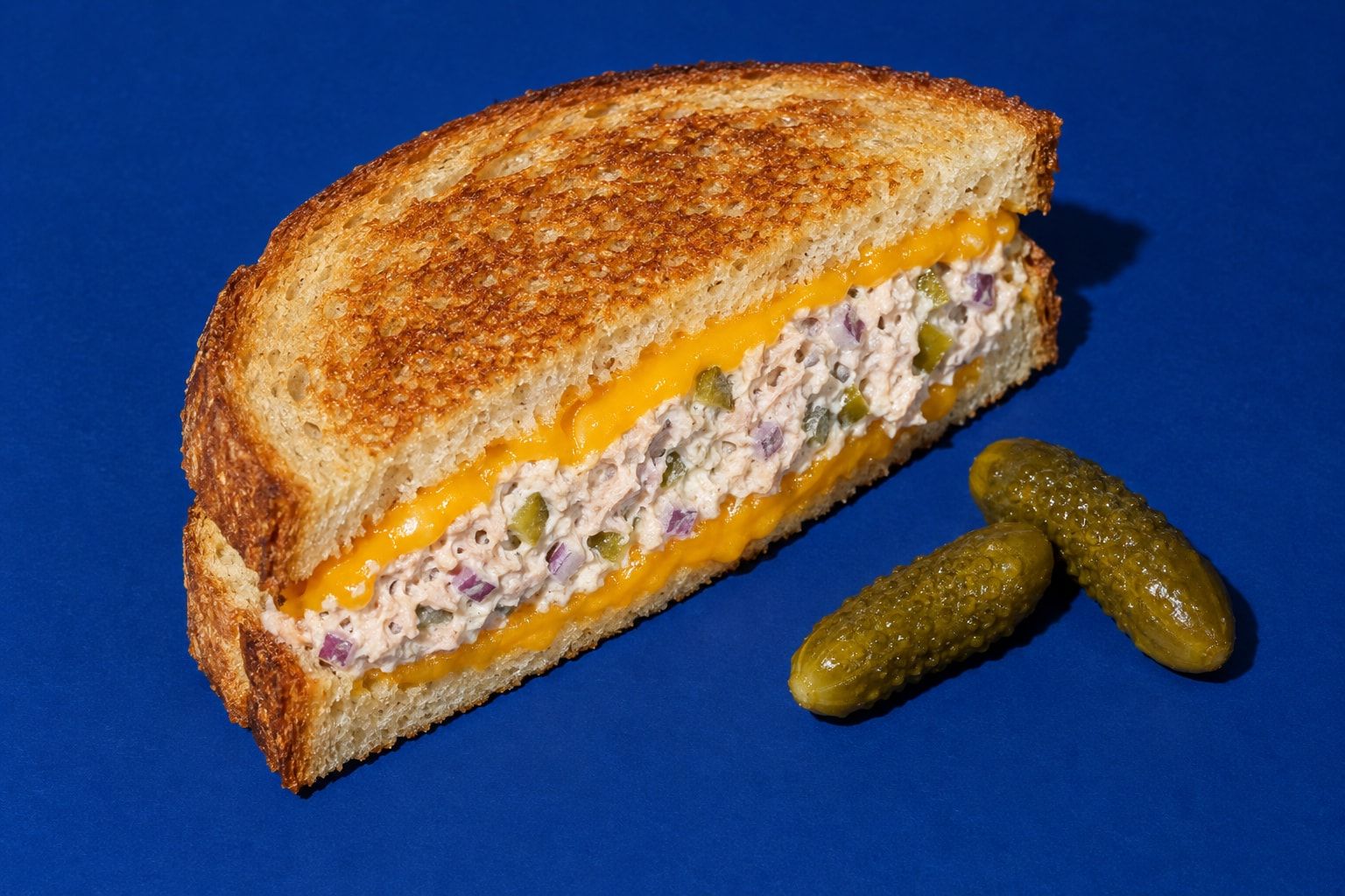 En delad tonfisktoast där snittytan visar krämig tonfiskröra med hackad saltgurka och rödlök mellan två lager smält cheddar.